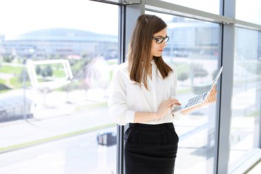 Dizüstü bilgisayarı olan güzel bir kadın uzman modern ofiste duruyor ve büyüleyici bir şekilde gülümsüyor. Tasarım, veri analizi, plan stratejisi üzerinde çalışıyorum. İş adamları konsepti.