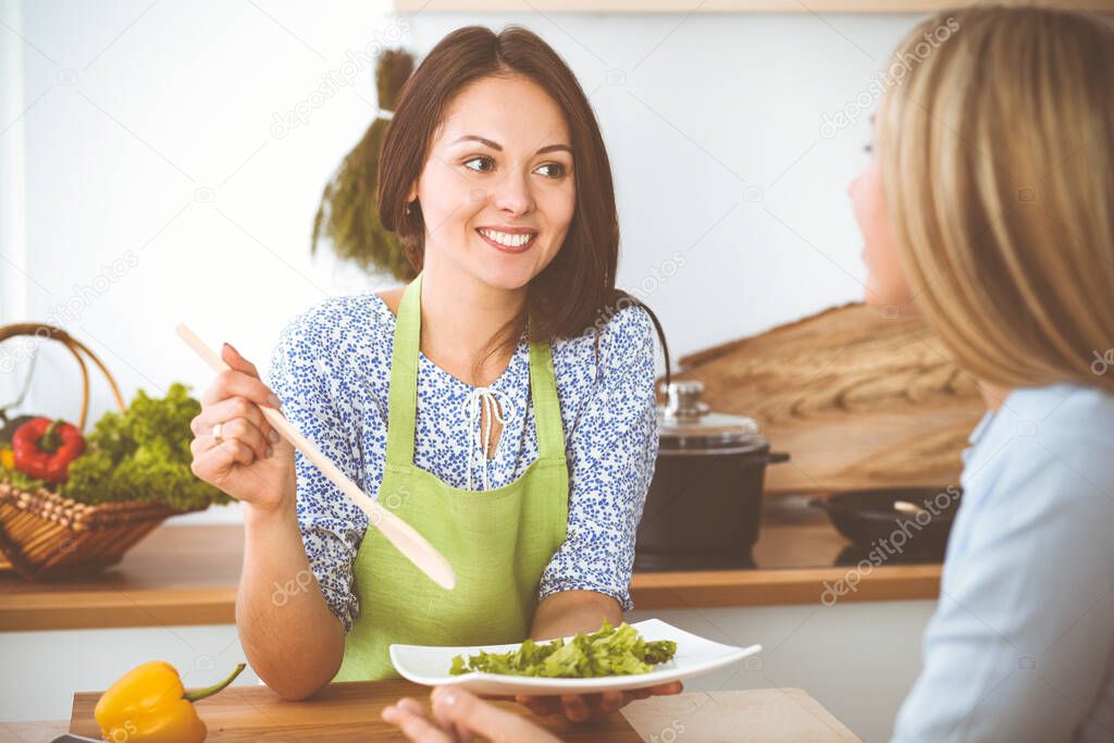Two women friends taste new recipes for delicious fresh salads while sitting at the kitchen table. Vegetarian cooking concept.