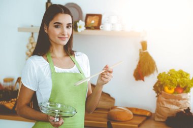 Çekici, genç, siyah saçlı bir kadın, güneşli mutfakta ayakta ve gülümserken tahta kaşıkla sıcak yemek tadıyor. Yemek pişirme ve ev idaresi kavramları.