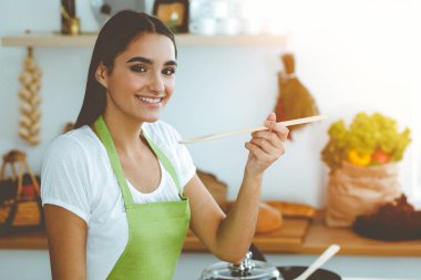 Çekici, genç, siyah saçlı bir kadın, güneşli mutfakta ayakta ve gülümserken tahta kaşıkla sıcak yemek tadıyor. Yemek pişirme ve ev idaresi kavramları.