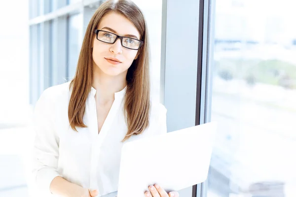 Beautiful female specialist with laptop computer standing in modern office and smiling charmingly. Working on design, data analysis, plan strategy. Business people concept.