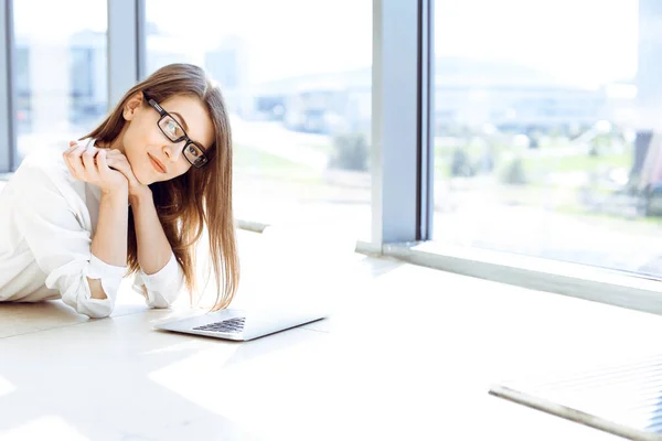 Beautiful female specialist with laptop computer laying on the floor in modern office and smiling charmingly. Working on design, data analysis, plan strategy. Business people concept.