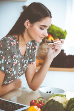 Çekici, genç, koyu saçlı bir kadın mutfakta masada otururken lezzetli bir salata karışımı için yeni bir tarif tadıyor. Tablet pc en iyi yemek kitabıdır.