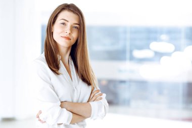 Kollarını kavuşturup modern ofiste duran ve büyüleyici bir şekilde gülümseyen güzel bir kadın uzman. Tasarım, veri analizi, plan stratejisi üzerinde çalışıyorum. İş adamları konsepti.