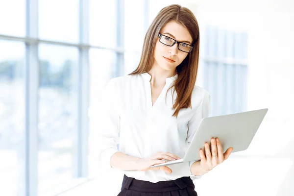 Beautiful female specialist with laptop computer standing in modern office and smiling charmingly. Working on design, data analysis, plan strategy. Business people concept.