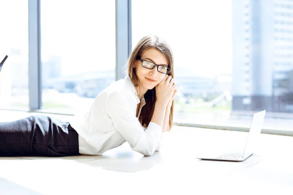 Beautiful female specialist with laptop computer laying on the floor in modern office and smiling charmingly. Working on design, data analysis, plan strategy. Business people concept.