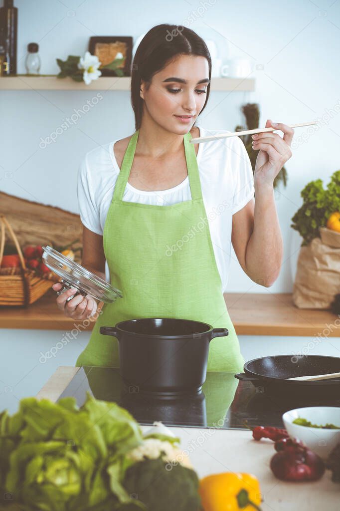 An attractive young dark-haired woman preparing soup by new keto recipe while standing and smiling in the kitchen. Cooking and householding concepts.