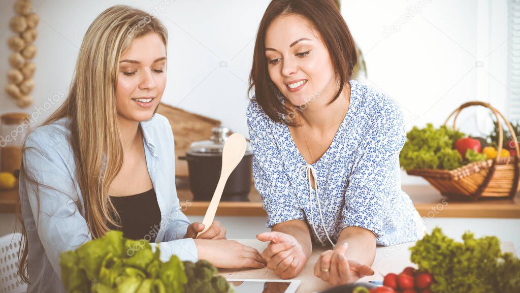Two women friends choosing the recipe for a delicious meal while sitting at the table in the kitchen. Tablet pc is the best cookbook.