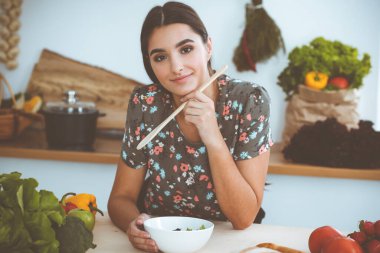 Çekici, genç, koyu saçlı bir kadın mutfakta oturup gülümserken lezzetli bir salata karışımı için yeni bir tarif tadıyor. Yemek pişirme ve ev idaresi kavramları.