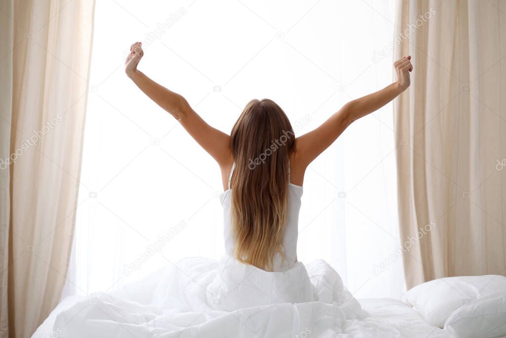 Woman stretching in bed after wake up, back view, entering a day happy and relaxed after good night sleep. Sweet dreams, good morning, new day, weekend, holidays concept.