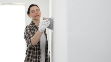 Koruyucu eldiven giyen ve kareli gömlekli bir kadın beyaz duvara macun yapıştırıyor, ev tadilatı sırasında spatulayla bileşik yayıyor, pratik ev geliştirme becerileri sergiliyor..