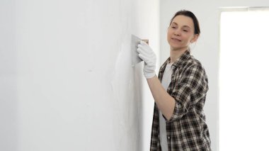 Koruyucu eldiven giyen ve kareli gömlekli bir kadın beyaz duvara macun yapıştırıyor, ev tadilatı sırasında spatulayla bileşik yayıyor, pratik ev geliştirme becerileri sergiliyor..