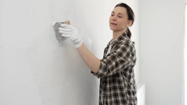 Koruyucu eldiven giyen ve kareli gömlekli bir kadın beyaz duvara macun yapıştırıyor, ev tadilatı sırasında spatulayla bileşik yayıyor, pratik ev geliştirme becerileri sergiliyor..