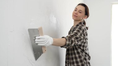 Kareli gömlek giyen kadın inşaat işçisi, beyaz duvar yüzeyini düzleştiriyor, profesyonel spatula tekniği ile macunu uyguluyor, iç dekorasyon konusunda yetenekli..