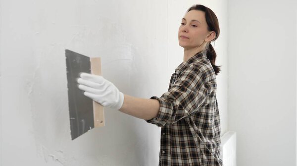 Woman construction professional wearing checkered shirt is smoothing white wall surface, applying putty with professional spatula technique, showcasing skilled interior renovation work.
