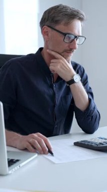 Professional businessman calculating financial data, reviewing documents near laptop in modern workplace setting. Audit and taxes, business concept