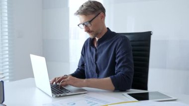 Focused businessman wearing glasses concentrating on his work, diligently typing on a laptop in a bright, modern office setting with a tablet and paperwork nearby. Business people concept