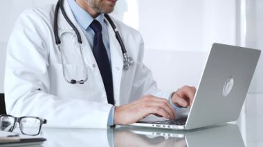 Steteskoplu beyaz önlüklü bir doktor laptopta yazı yazıyor. Teletıp, sağlık ve dijital tıbbi kayıtları temsil ediyor. Tıp, sağlık ve bilim konsepti