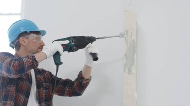 Kırmızı kareli gömlek, mavi başlık ve koruyucu eldiven giyen inşaat işçisi, döner çekiç matkabıyla beyaz duvarı yıkıyor, toz ve portre görüntüsü yaratıyor. Yenileme konsepti.