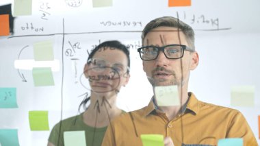 İş arkadaşları beyin fırtınası yapıyor, cam tahtaya strateji notları yazıyor, yaratıcı iş yeri toplantısında renkli yapışkan notlarla işbirliği yapıyorlar. Takım çalışması konsepti.