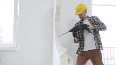 Bej kareli gömlek, sarı başlık ve koruyucu eldiven giyen erkek inşaat işçisi, dönen çekiç matkabıyla beyaz duvarı yıkıyor ve orta uzunlukta toz üretiyor. Yenileme projesi