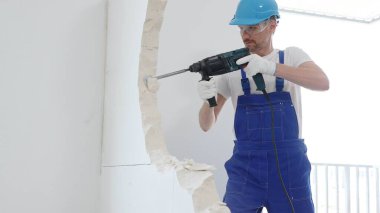 Mavi tulumlu ve koruyucu beyaz eldivenli bir inşaat işçisi, beyaz duvarı döner çekiç matkabıyla yıkıyor, toz üretiyor, yatay görüşü kapatıyor. Yenileme konsepti.