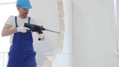 Mavi tulumlu, koruyucu şapkalı ve beyaz eldivenli bir inşaat işçisinin portresi, döner çekiç matkabıyla beyaz duvarı yıkıyor, toz oluşturuyor, yatay görüş sağlıyor. Yenileme.