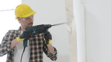 Bej kareli gömlek, sarı başlık ve koruyucu eldiven giyen bir inşaat işçisinin portresi, döner çekiç matkabıyla beyaz duvarı yıkıyor ve toz oluşturuyor. Yenileme konsepti.