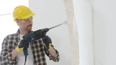 Bej kareli gömlek, sarı başlık ve koruyucu eldiven giyen bir inşaat işçisinin portresi, döner çekiç matkabıyla beyaz duvarı yıkıyor ve toz oluşturuyor. Yenileme konsepti.