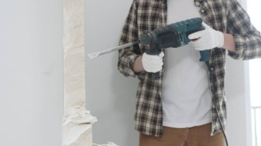Tanınmayan bej kareli gömlek ve koruyucu eldivenler giyen erkek inşaat işçisi, döner çekiç matkabıyla beyaz duvarı yıkıyor, toz üretiyor, yakın görüş sağlıyor. Yenileme konsepti.