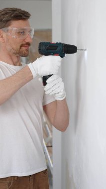 Beyaz tişört ve koruyucu eldiven giyen orta yaşlı erkek işçi, ev yenileme çalışmaları sırasında kablosuz matkapla duvarda delik açıyor. Dikey görünüm.