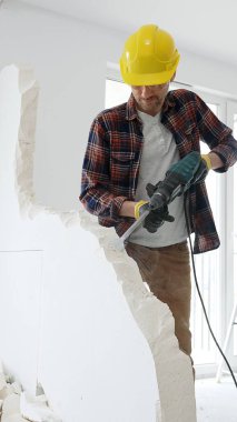 Kırmızı kareli gömlek, sarı başlık ve koruyucu eldiven giyen erkek inşaat işçisi, dönen çekiç matkabıyla beyaz duvarı yıkıyor ve orta uzunlukta toz üretiyor. Yenileme projesi.