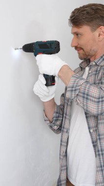 Gri gömlekli ve beyaz eldivenli orta yaşlı profesyonel işçi, ev yenileme çalışmaları sırasında kablosuz matkapla duvarda delik açıyor. Dikey portre görünümü.