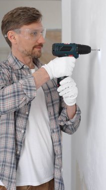 Gri gömlekli ve beyaz eldivenli orta yaşlı işçi, ev yenileme çalışmaları sırasında kablosuz matkapla duvarda delik açıyor. Dikey görünüm.