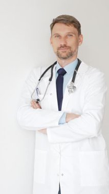 Kendine güvenen erkek doktor kolları çapraz duruyor, steteskop ve beyaz önlük giyiyor. Düşünceli bir şekilde, tıp alanında profesyonelliği ve uzmanlığı somutlaştırıyor. İlaç ve sağlık hizmetleri