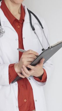 Tuğla kırmızı bluzun üzerine steteskop ve beyaz önlük giyen kimliği belirsiz kadın doktor tıbbi uzmanlığı ve hasta bakımını temsil eden bir panoya yazı yazıyor. İlaç ve sağlık hizmetleri kavramı.