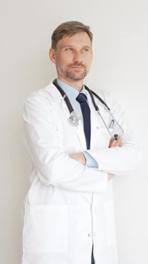 Kendine güvenen erkek doktor kolları çapraz duruyor, steteskop ve beyaz önlük giyiyor. Düşünceli bir şekilde, tıp alanında profesyonelliği ve uzmanlığı somutlaştırıyor. İlaç ve sağlık hizmetleri