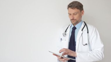 Beyaz önlüklü ve steteskoplu bir doktor dijital tablet kullanıyor. Hasta kayıtlarını güncellemeye odaklanmış, modern sağlık teknolojisi ve profesyonel uzmanlığın altını çizmiş. İlaç ve sağlık hizmetleri