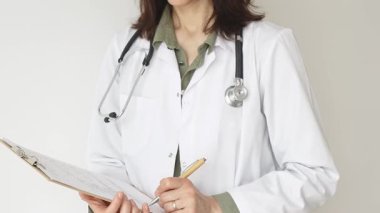 Stetoskop ve yeşil bluzun üzerine beyaz önlük giyen kimliği belirsiz bir kadın doktor tahta renkli bir panoya tıbbi uzmanlığı ve hasta bakımını temsil eden bir yazı yazıyor. Tıp konsepti