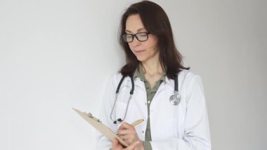 Yeşil bluzun üzerine steteskop, siyah gözlük ve beyaz önlük takan doktor kadın tıbbi uzmanlığı ve hasta bakımını temsil eden bir panoya yazı yazıyor. İlaç ve sağlık hizmetleri