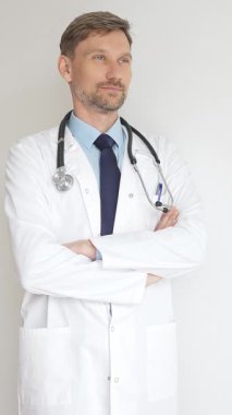 Kendine güvenen erkek doktor kolları çapraz duruyor, steteskop ve beyaz önlük giyiyor. Düşünceli bir şekilde, tıp alanında profesyonelliği ve uzmanlığı somutlaştırıyor. İlaç ve sağlık hizmetleri