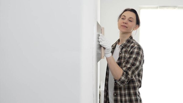 Female construction professional wearing checkered shirt is smoothing white wall surface, applying putty with professional spatula technique, showcasing skilled interior renovation work.