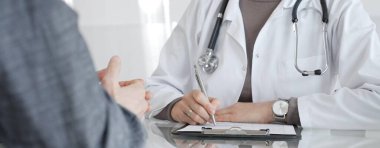 Klinikteki hasta muayenesi sırasında panoya not alan beyaz önlüklü doktor sağlık, teşhis ve bakım kararlarına odaklanmış. İlaç ve sağlık hizmetleri