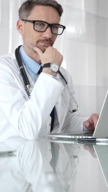 Kendine güvenen beyaz önlüklü erkek doktor laptopta çalışıyor, tıbbi verileri analiz ediyor ve modern hastane ortamında hasta bakımını planlıyor. İlaç ve sağlık hizmetleri