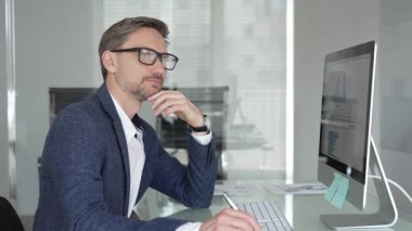 Profesyonel iş adamı gözlük takıyor ve spor ceket giyiyor, çağdaş bir ofis ortamında çalışıyor, bir masaüstü bilgisayarında görevler yapıyor. İş adamları konsepti