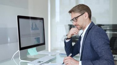 Profesyonel iş adamı gözlük takıyor ve spor ceket giyiyor, çağdaş bir ofis ortamında çalışıyor, bir masaüstü bilgisayarında görevler yapıyor. İş adamları konsepti