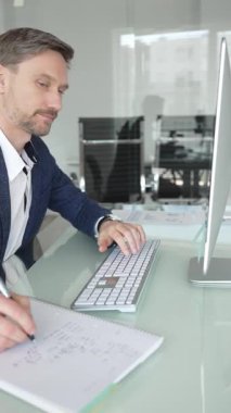 Takım elbiseli işadamı masa başına odaklanmış, klavyeye yazarken aynı zamanda kalemle not alıyor, modern ofis ortamında birden fazla iş yapıyor. İş adamları konsepti