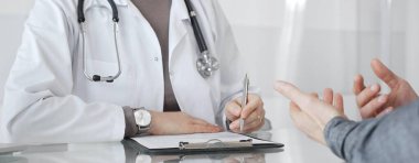 Klinikteki hasta muayenesi sırasında panoya not alan beyaz önlüklü doktor sağlık, teşhis ve bakım kararlarına odaklanmış. İlaç ve sağlık hizmetleri