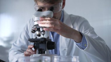 Laboratuvar önlüğü giyen ve koruyucu gözlük takan erkek bilim adamı mikroskop kullanarak modern laboratuvar düzeninde araştırma yapıyor, portre görüntüsü çiziyor. Gösterişsiz ışıklandırma fotoğrafı. Bilim ve Tıp Konsepti.