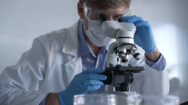 Mavi eldiven, koruyucu gözlük ve tıbbi maske takan erkek bilim adamı araştırmacı modern laboratuarda mikroskop kullanıyor, portre görüşü. Gösterişsiz ışıklandırma fotoğrafı. Bilim ve Tıp.